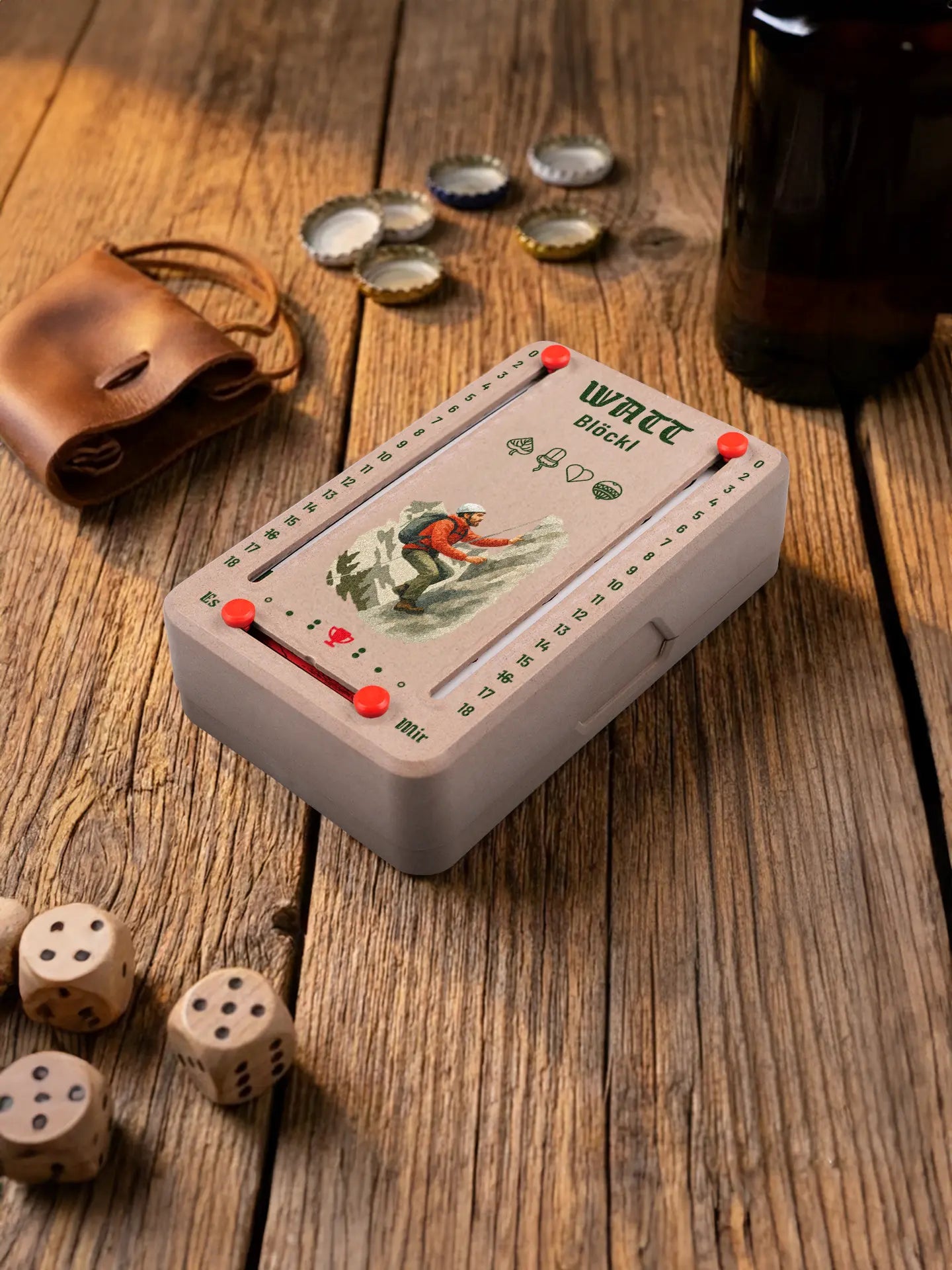 Wattblöckl in Holzoptik mit roten Pins und Bergsteiger-Motiv. Platziert auf Holztisch mit Würfeln und Kronkorken.